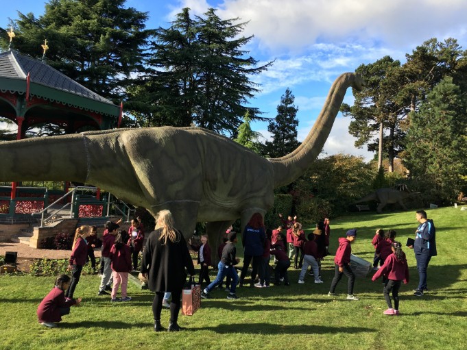 Des enfants rassemblés autour d'un grand dinosaure animatronique au long cou à Récréo Parc à Montréal, avec des arbres et les terrains du parc visibles en arrière-plan