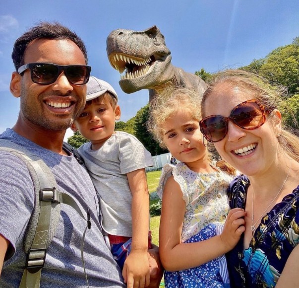 A smiling family of four posing outdoors with a life-sized animatronic T-Rex dinosaur against a bright blue sky and green hillside during summer 2026