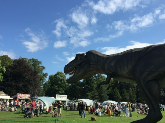 Life-sized animatronic T. rex towering over families at Jurassic Kingdom outdoor dinosaur exhibition in a sunny Canadian park